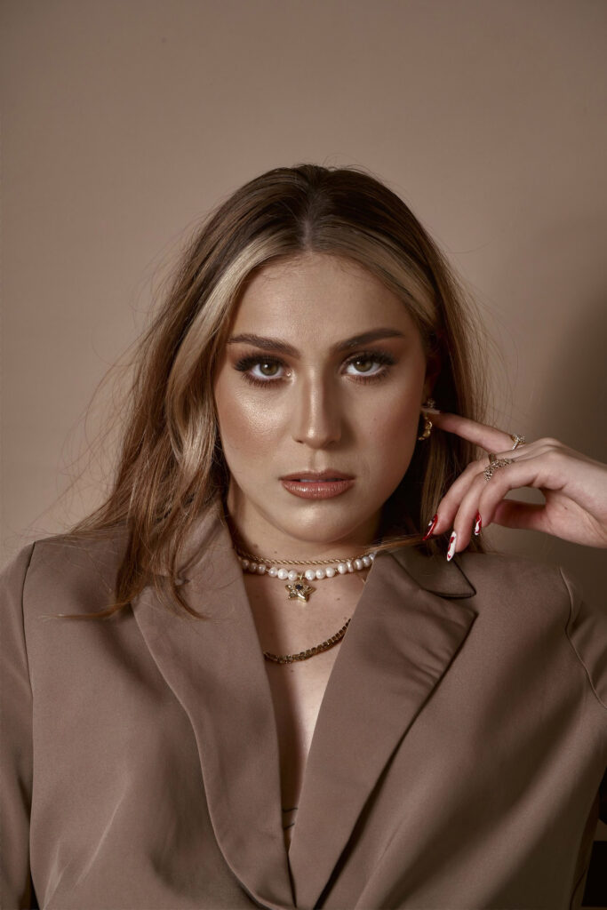 Clarisa Cisneros a model with striking features poses against a muted background showcasing her makeup jewelry and attire.